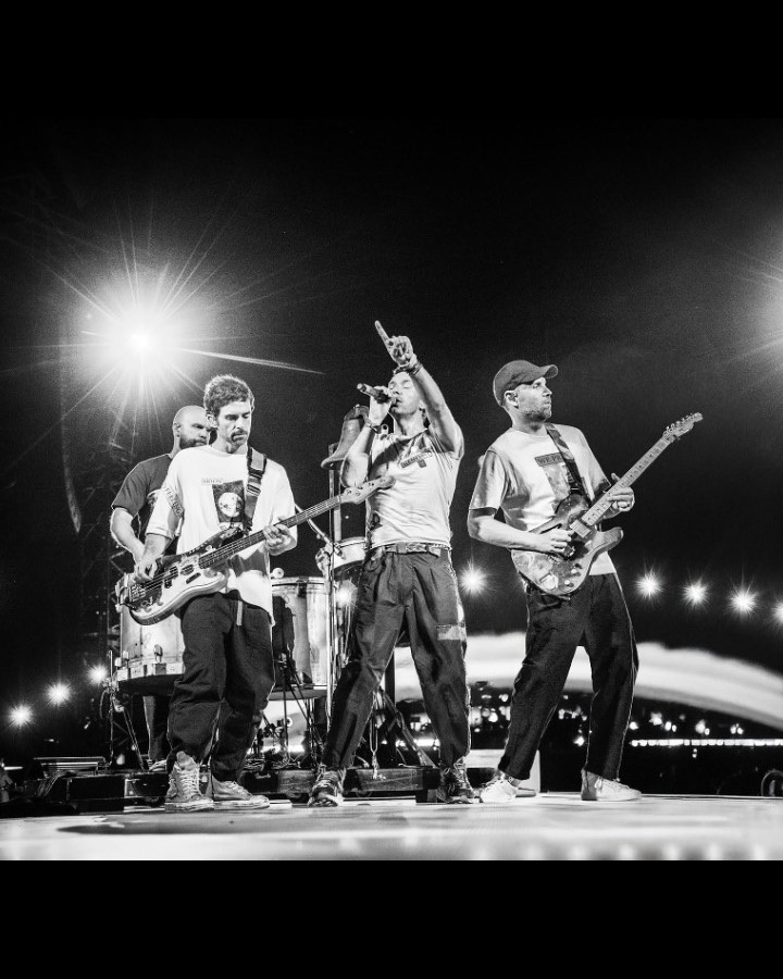 Black and white photograph of Coldplay performing on stage, Chris Martin pointing upward under stadium lights